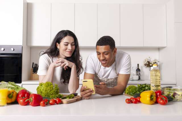 A smiling couple looks at a smartphone while unpacking healthy groceries in their kitchen.