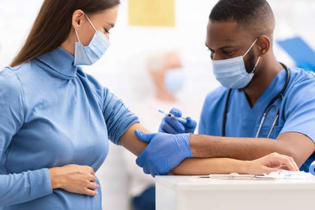  A pregnant woman in a light blue turtleneck and mask receives a vaccine or injection in her arm from a male nurse in blue scrubs, mask, and gloves.