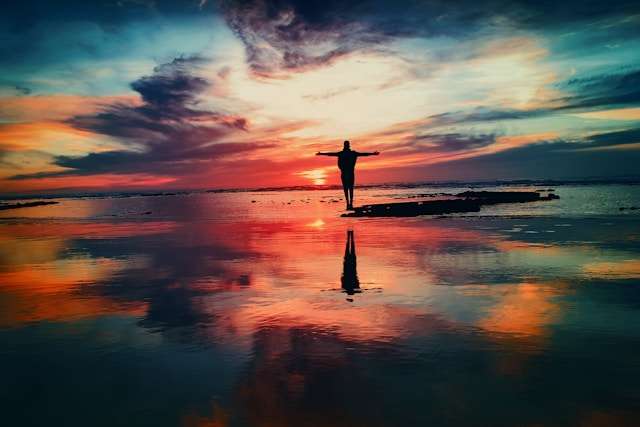 Silhouette of a person with outstretched arms against a colorful sunset reflection.