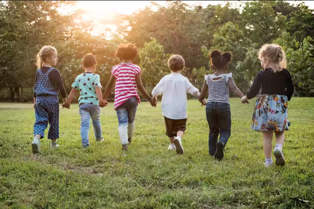 Six children of diverse backgrounds walk hand-in-hand across a grassy field towards a setting sun.