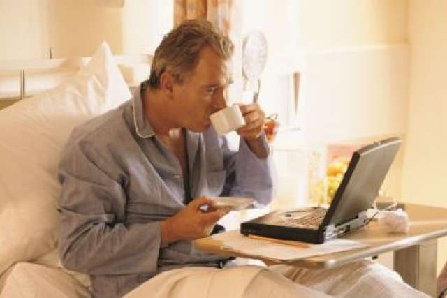 Man drinking tea in bed while using laptop and holding phone and newspaper.
