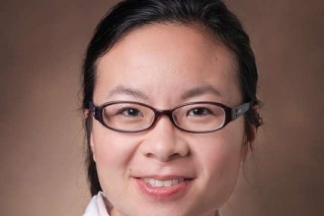 Smiling person in glasses and white lab coat against warm brown background.