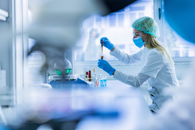 Researcher inserting liquid into vial with syringe, wearing a mask in a laboratory.