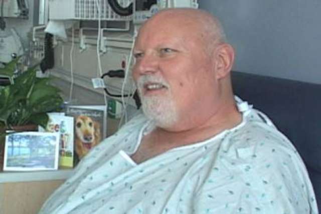 Alan Landros in hospital gown smiling at bedside.