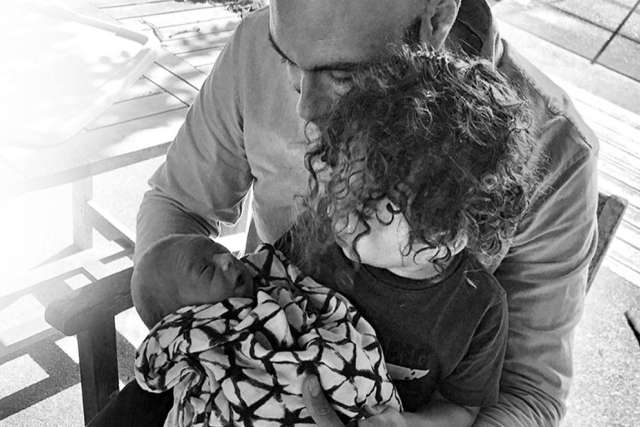 Black and white photo: An adult man holds a swaddled newborn baby while being embraced by a young child with curly hair in a bright, outdoor setting.