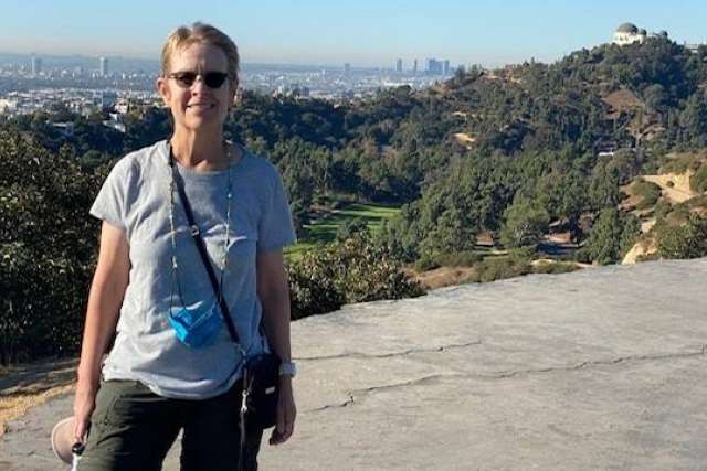 Laurie Adami, a woman with short hair and sunglasses, smiles confidently outdoors with a city skyline and hills in the background.