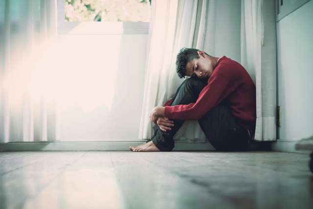 A dejected young man sits crouched in a corner with his arms wrapped around his legs and his head resting on his knees.