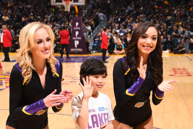 Gio, a child, stands on a basketball court between two cheerleaders in black and gold outfits, clapping their hands and smiling.