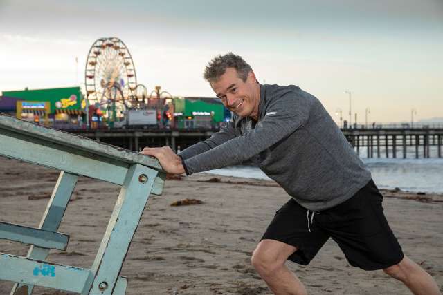 Man pushing a lifeguard stand on the beach with a pier and ferris wheel in the background.