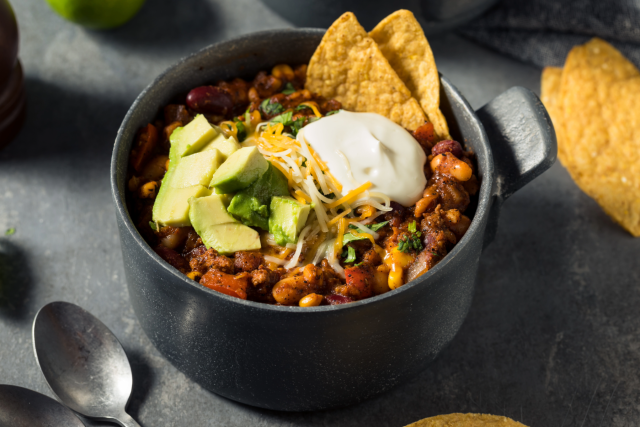 Bowl of chili topped with avocado slices, sour cream, cilantro, and tortilla chips