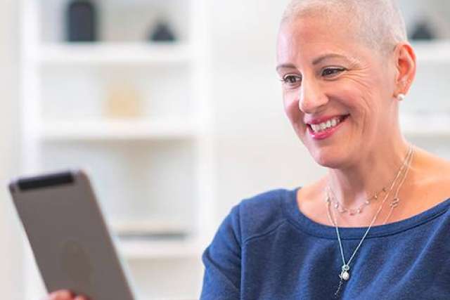  A smiling woman with a shaved head, wearing a navy blue top and necklaces, looks down at a tablet she is holding in an indoor, well-lit setting.