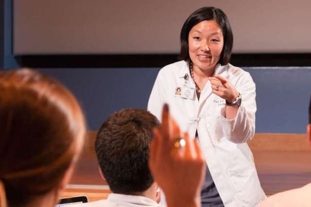 A person in a lab coat speaks to an audience in a classroom setting.