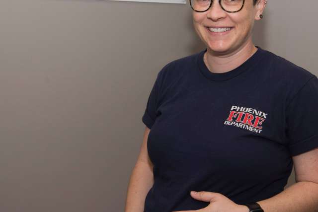 Person wearing a dark T-shirt with “Phoenix Fire Department” logo, standing against a plain wall with hands visible.