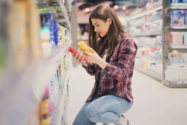 Person wearing a plaid shirt and jeans kneeling in a store aisle, comparing two bottles of products with shelves of items in the background.
