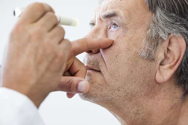 Older man receiving an eye exam as a clinician shines a light into his eye to check vision and pupil response.