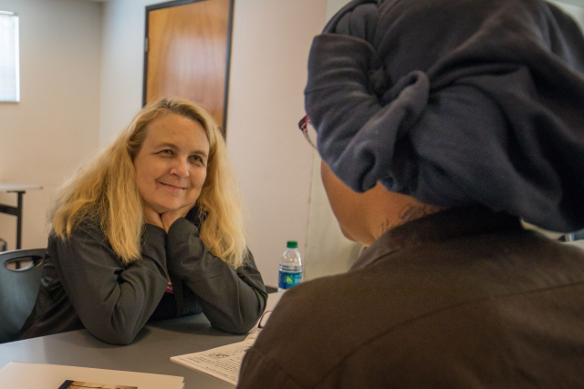 A nurse and another woman converse, sitting across a table from each other.