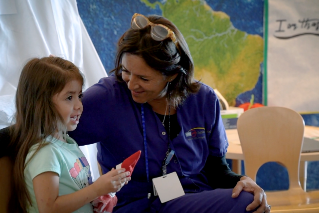 Child Life specialist in scrubs smiling and sitting beside a young girl holding a toy in a hospital playroom.