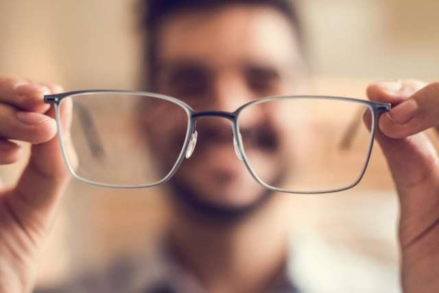 Hands holding eyeglasses toward camera, blurred face behind