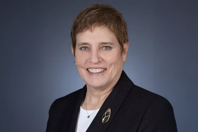 Professional headshot of a person wearing a dark blazer against a dark background.