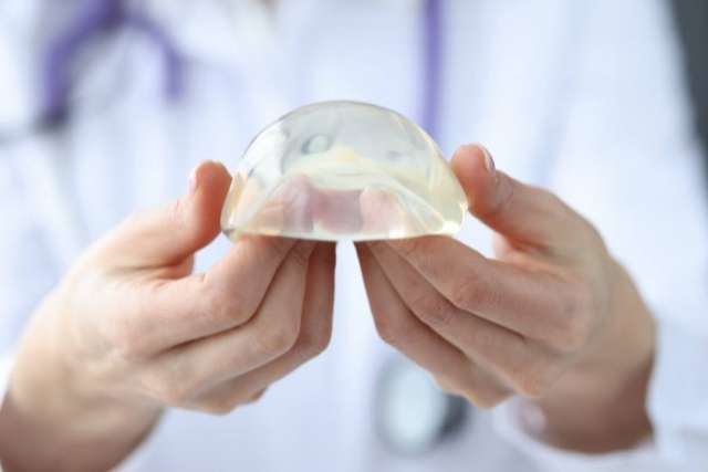 A person in a white lab coat holds up a clear, half-dome-shaped breast implant with both hands.
