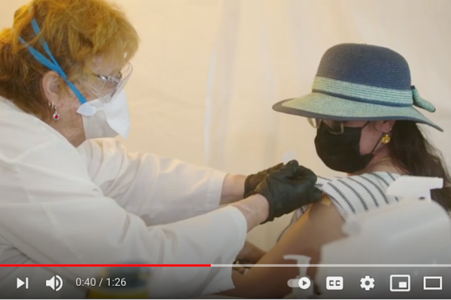 A medical professional wearing a mask and gloves administers a vaccine to a person. The person receiving the shot is wearing a hat and a mask.