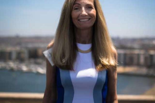 A smiling woman with long blonde hair, wearing a white and blue dress and gold necklace, stands outdoors with a city skyline visible.
