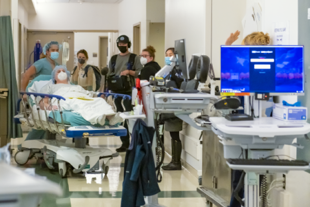 Chloe on a gurney is wheeled through a busy hospital hallway by medical staff, with others standing nearby.