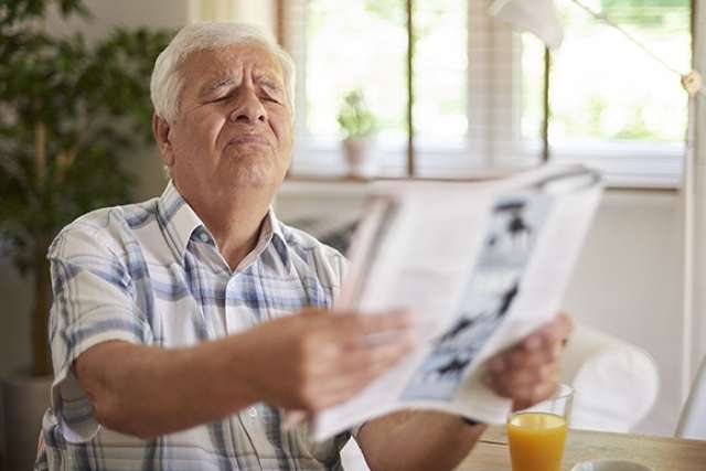 Older adult wearing a plaid shirt sitting at a table, holding and reading a newspaper with a glass of orange juice nearby.