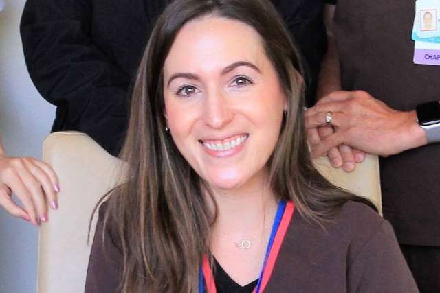 Person wearing a dark scrub top with a medal ribbon around the neck, seated on a light-colored chair with others nearby.