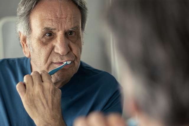 Older man brushing his teeth while looking in a bathroom mirror.