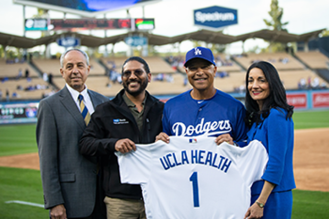 Four people pose at Dodger Stadium, holding a Dodgers jersey with “UCLA Health” and the number 1 on the back.