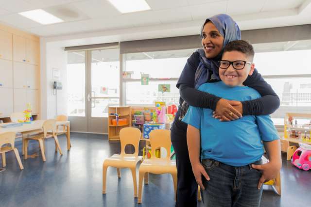 A woman holding a child around his chest lovingly inside of a classroom environment.