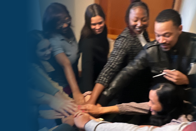 A group of individuals gathered in a circle each have one hand reaching toward center atop of each other