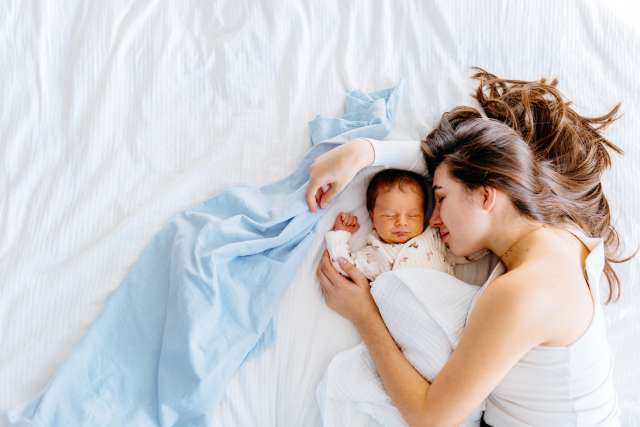 Woman lying in bed beside a newborn, gently holding the baby on white bedding