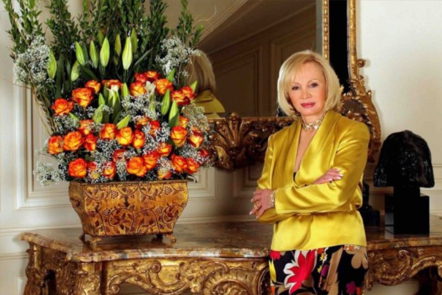 Iris Cantor in a yellow blouse poses beside a large flower arrangement and ornate mirror.