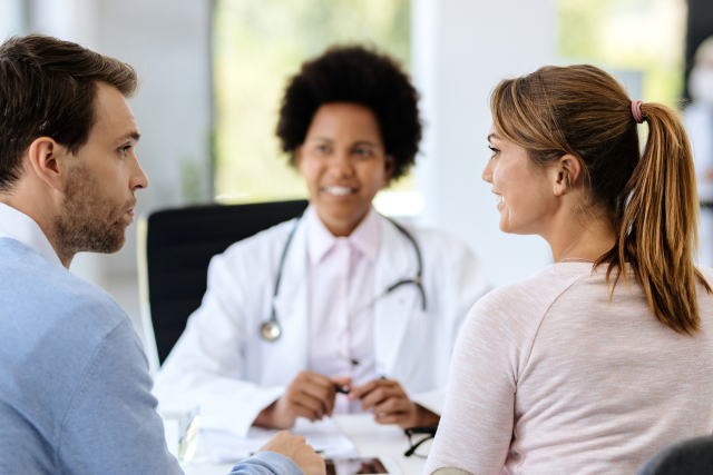 Doctor smiles while speaking with a couple during a consultation in a bright, modern medical office.