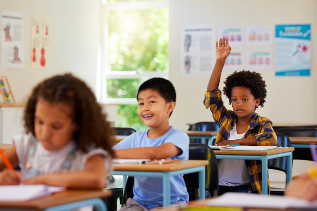 three children sitting in a classroom at their desks, with one child raising their hand