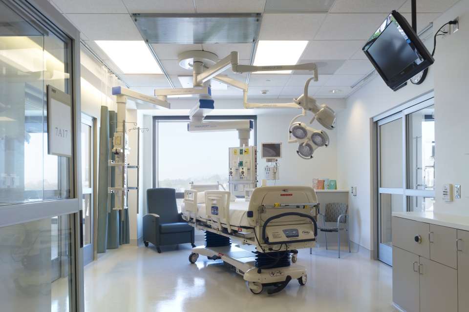 An image of a brightly lit Intensive Care Unit (ICU) room at Ronald Reagan UCLA Medical Center. The room contains a hospital bed, medical equipment, a monitor, and a blue armchair.