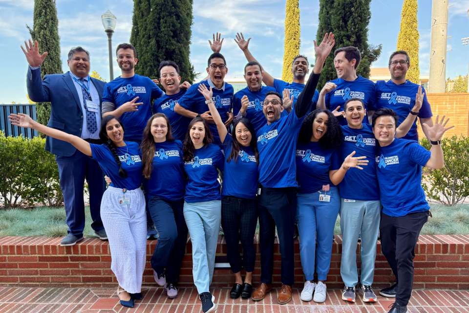 Group of people standing outside with their hands up in the air cheering with blue shirts in an outdoor setting.