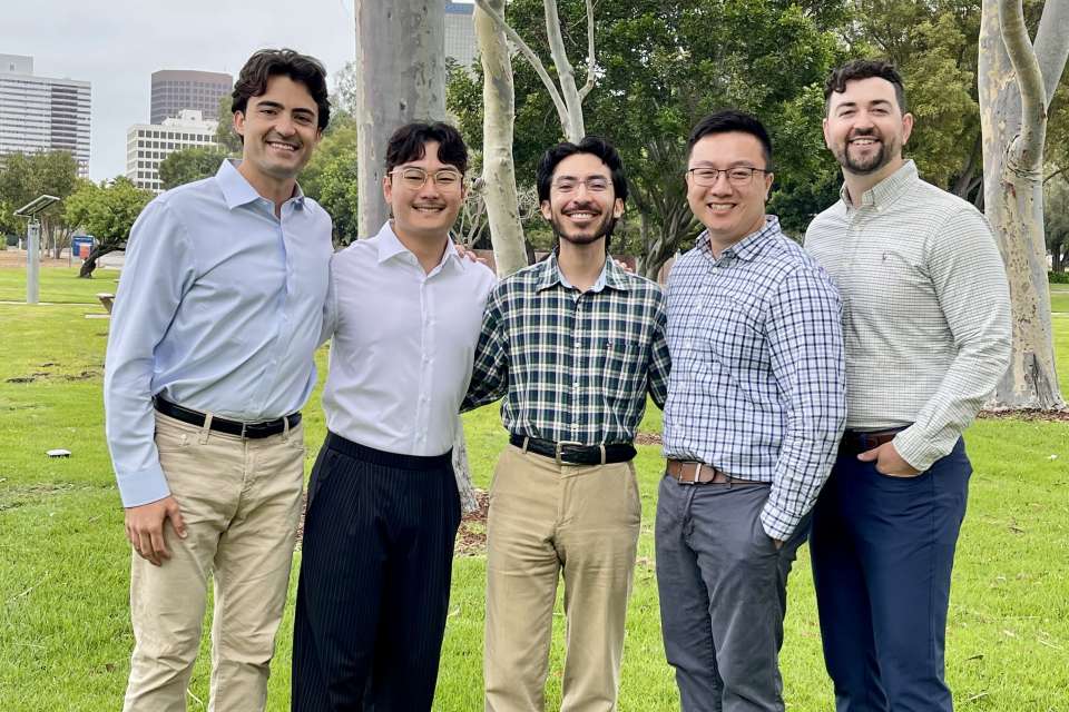 Five men standing together on a park lawn with trees and a city skyline behind them