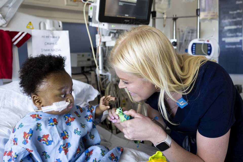 Blonde nurse showing a toy turtle to an infant that is sitting on a hospital bed