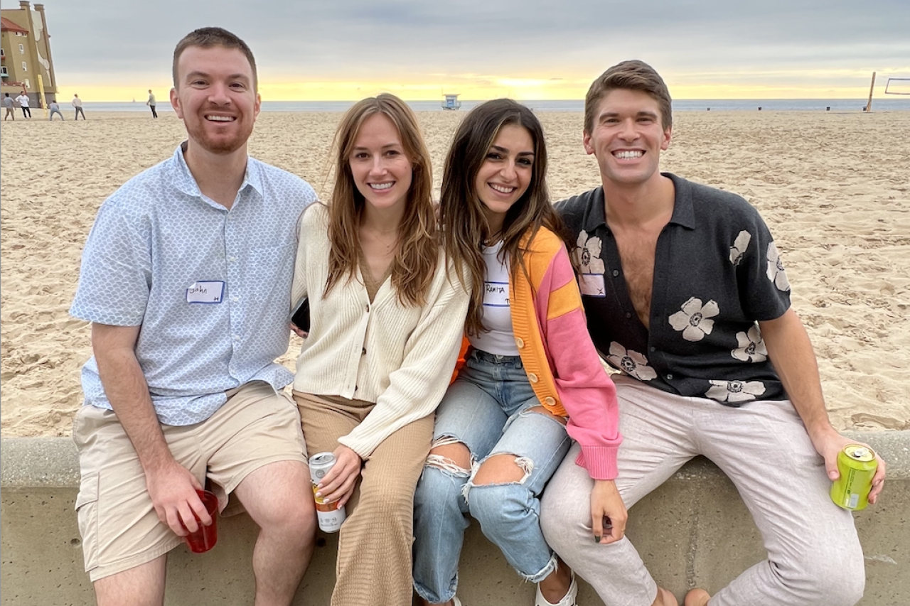 Four young adults, Chief Residents, at the DAPM resident retreat in 2022, sit on a low wall at the beach, casually dressed and holding drinks.