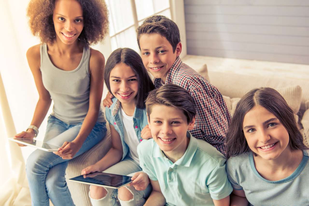 Five smiling kids sitting together on a couch. Two of them are holding tablets.