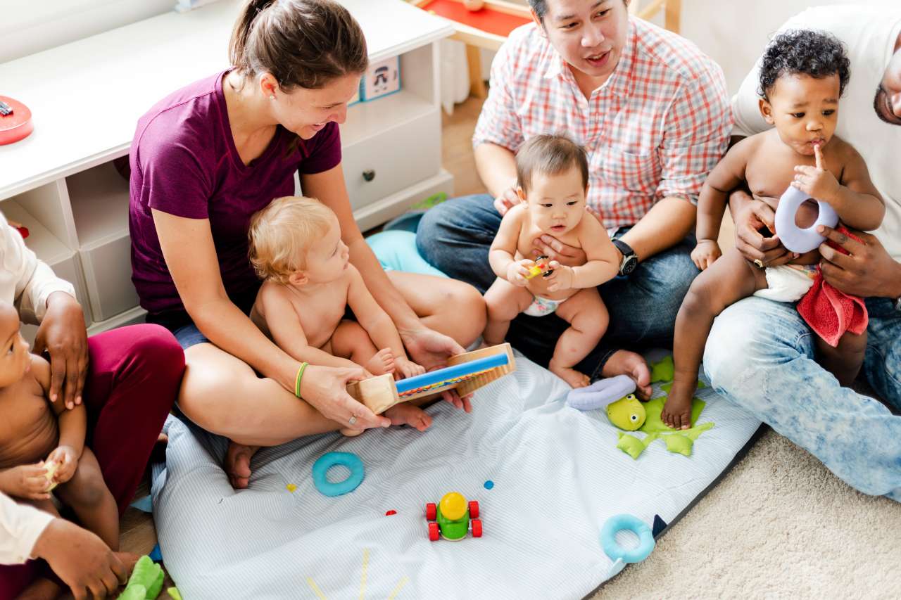 Babies and adults seated in a circle on a blanket sharing toys.