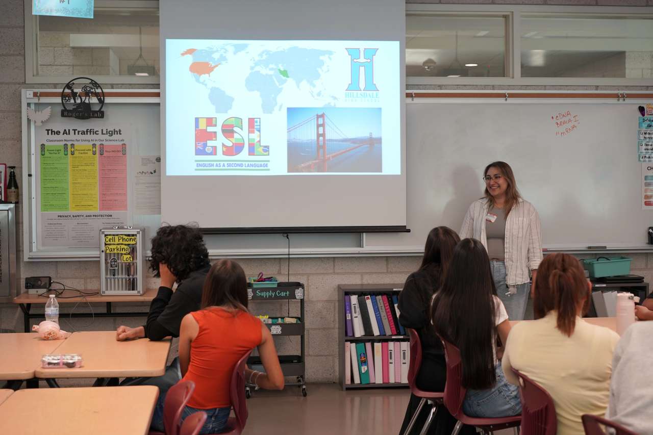 A woman speaks to a classroom of seated students beside a projected slide showing a world map with the US and Iran highlighted, an "ESL - English as a Second Language" flag graphic, a photo of the Golden Gate Bridge, and the Hillsdale High School logo.