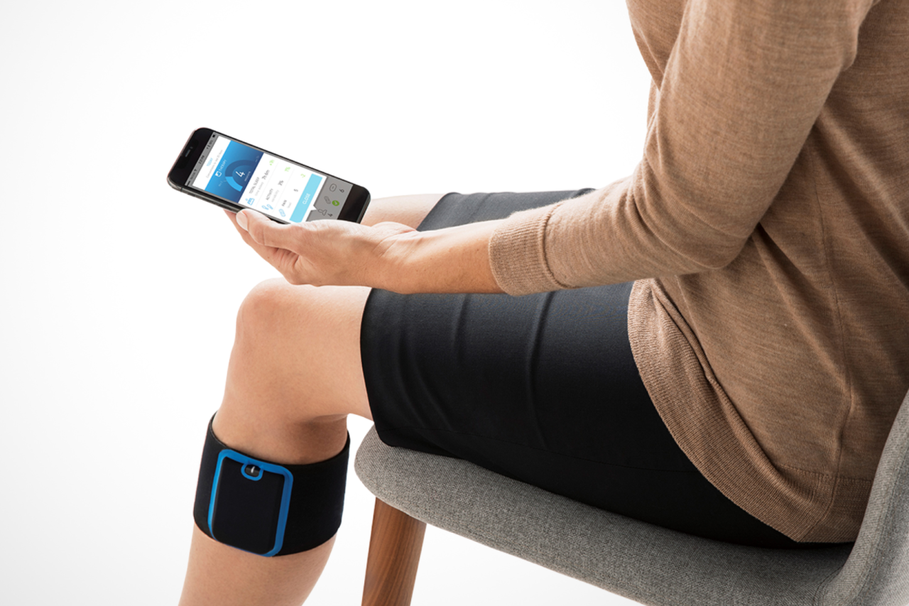 A person is seated with a Quell device strapped to their leg and holding a smartphone, managing the device.