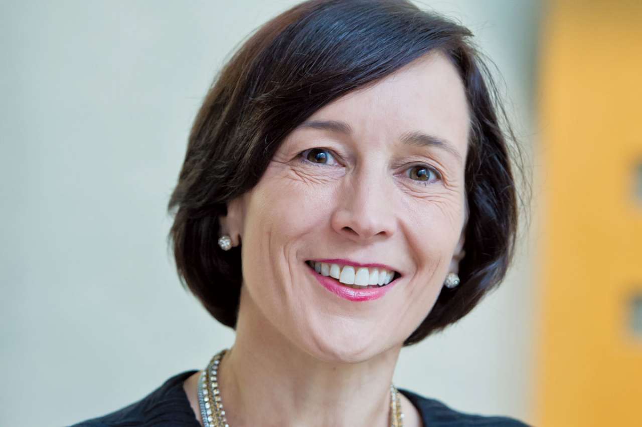 Woman wearing a black dress and layered necklaces, standing in a modern setting.