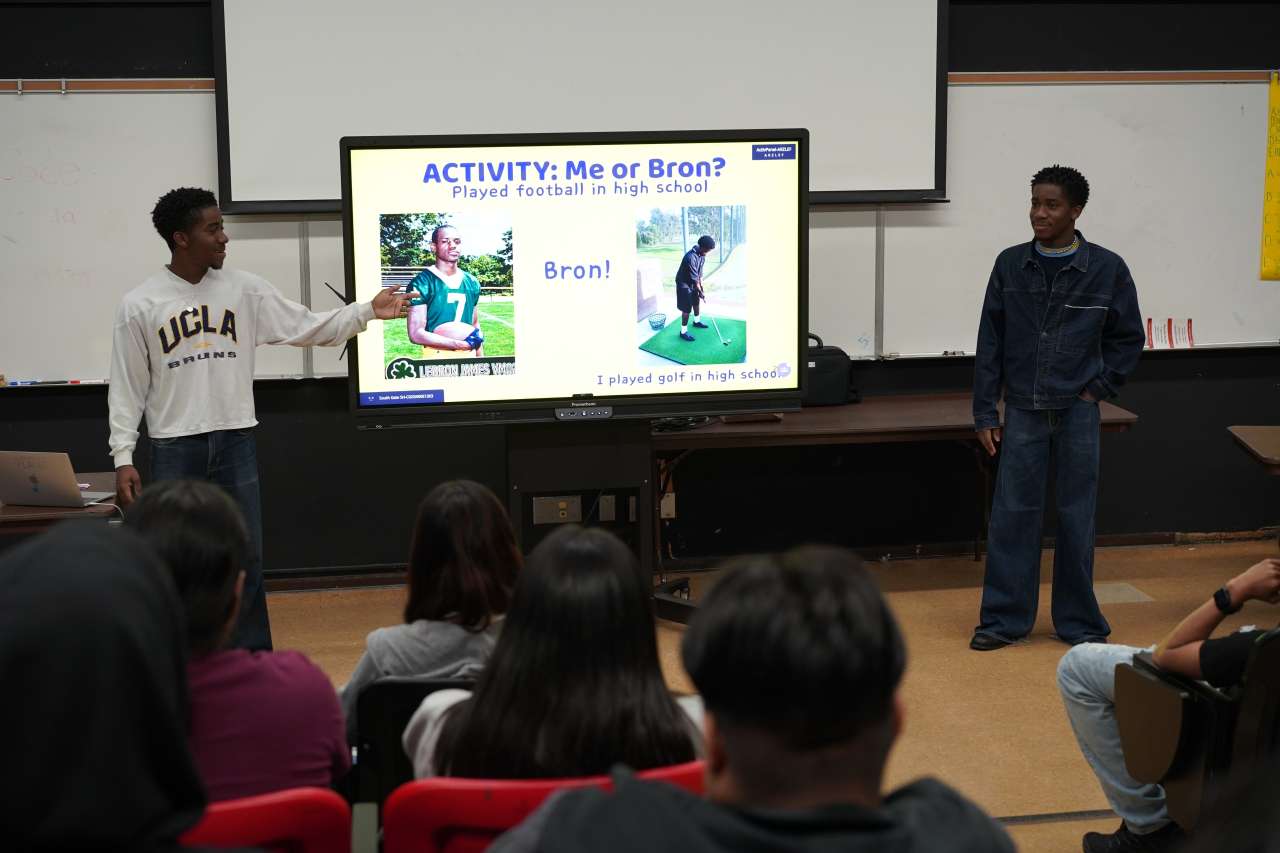 A student in a UCLA sweatshirt gestures toward a classroom presentation screen displaying "ACTIVITY: Me or Bron?" with a "Played football in high school" trivia slide, while another student stands to the right smiling. Classmates are seated in the foreground watching the presentation.