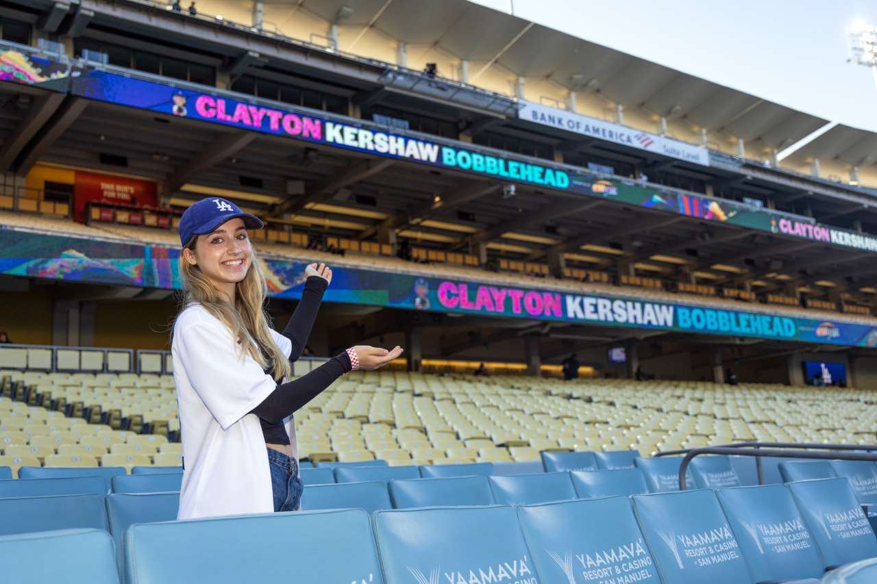 Josie Le Blanc is in the stands at Dodger Stadium gesturing to a sign that announces it's Clayton Kershaw bobblehead night.