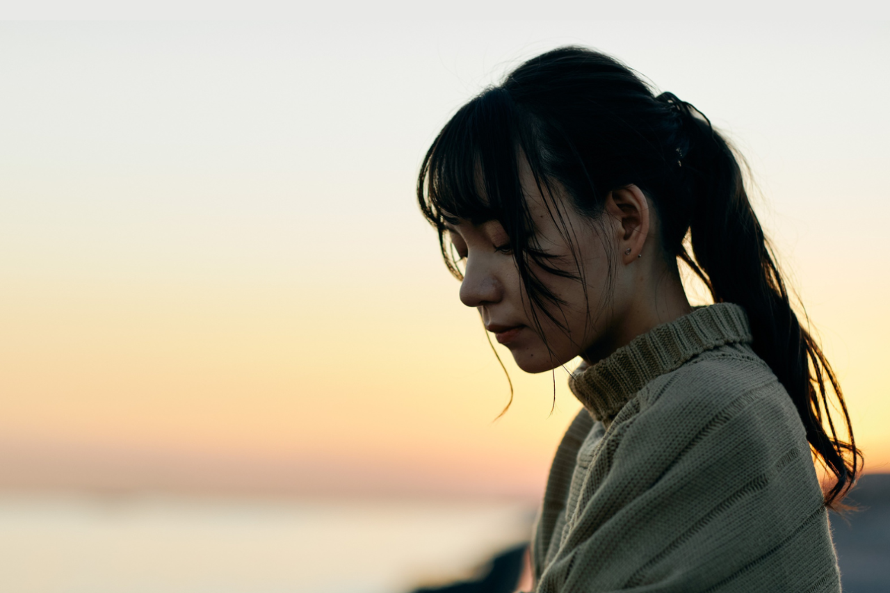 Woman in a beige sweater looking down thoughtfully at sunset by the water.
